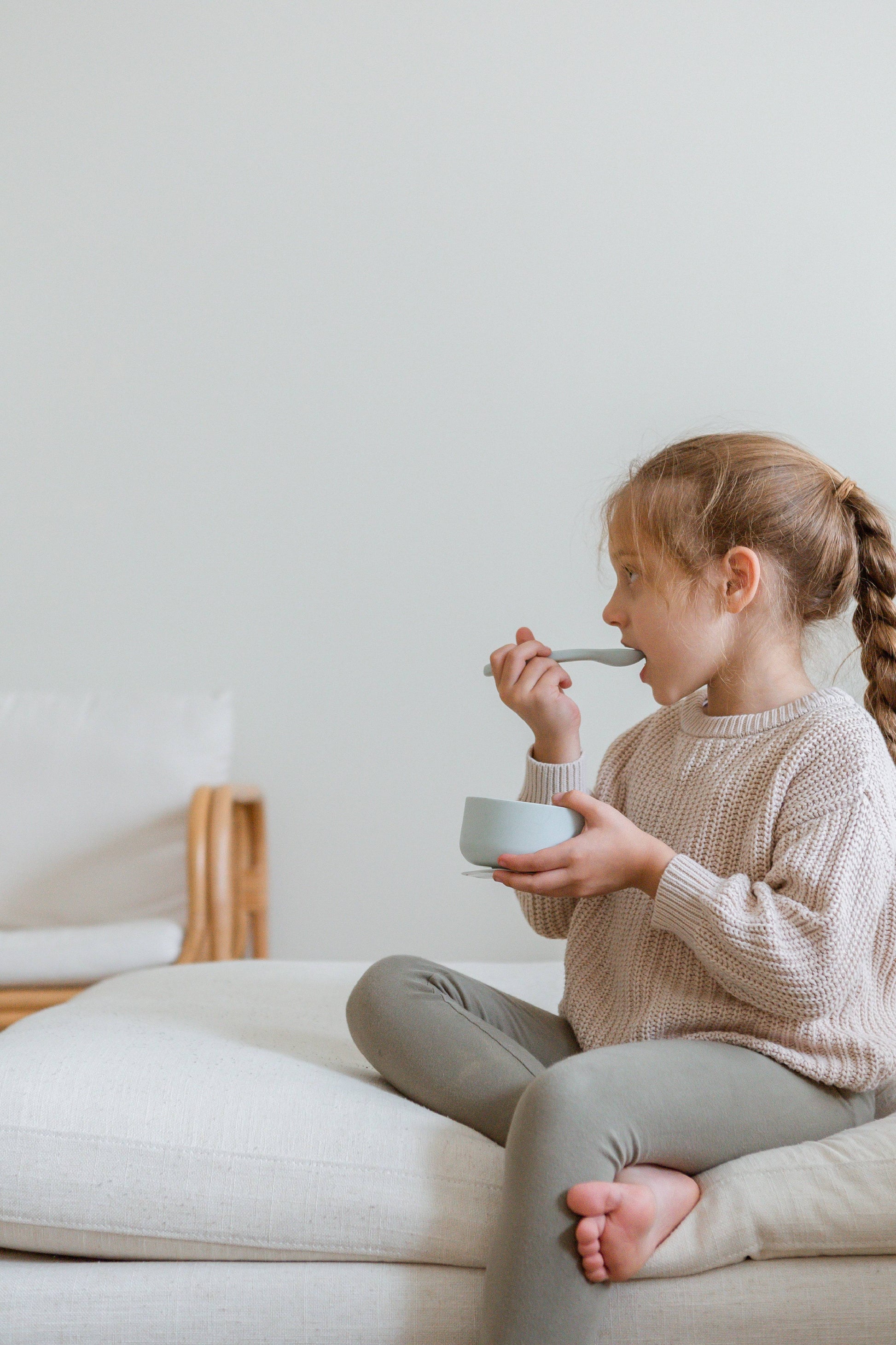 A young girl with a braid sits cross-legged on a light couch, wearing a beige sweater and gray leggings, feeding from a bowl using Soft Spot Soft Baby Spoons (set of 2) by Soft Spot against a plain white background.