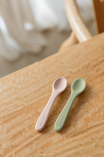 Two Soft Spot Soft Baby Spoons, designed for babys first gentle feeding, lie side by side on a light wooden table. The softly blurred background and part of a chair add to the serene scene. Brand: Soft Spot. Set of 2.