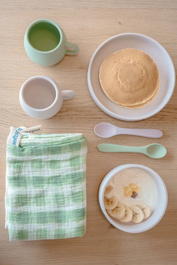 A tabletop features pancakes, a bowl of yogurt with banana slices and raisins, a pair of Soft Spot Soft Baby Spoons by Soft Spot—gentle on teething gums—a green cup, white cup, and a folded green plaid cloth.