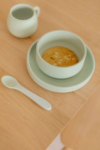 A light green bowl with a cookie sits on a matching plate next to the Soft Spot Soft Baby Spoons, Set of 2 by Soft Spot, and a small jug—perfect for easy feeding on a wooden table.