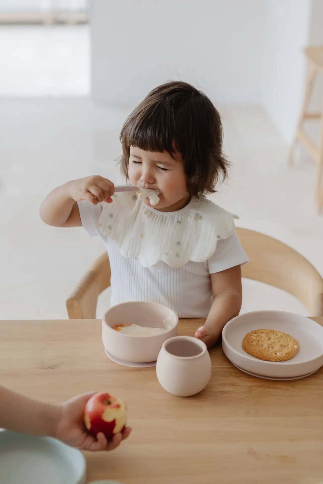 A young child with brown hair sits at a wooden table, enjoying feeding with Soft Spot Soft Baby Spoons. The child wears a white ruffled bib, and there’s a cookie, cup, and an apple in another hand in the foreground. Set of 2 by Soft Spot.