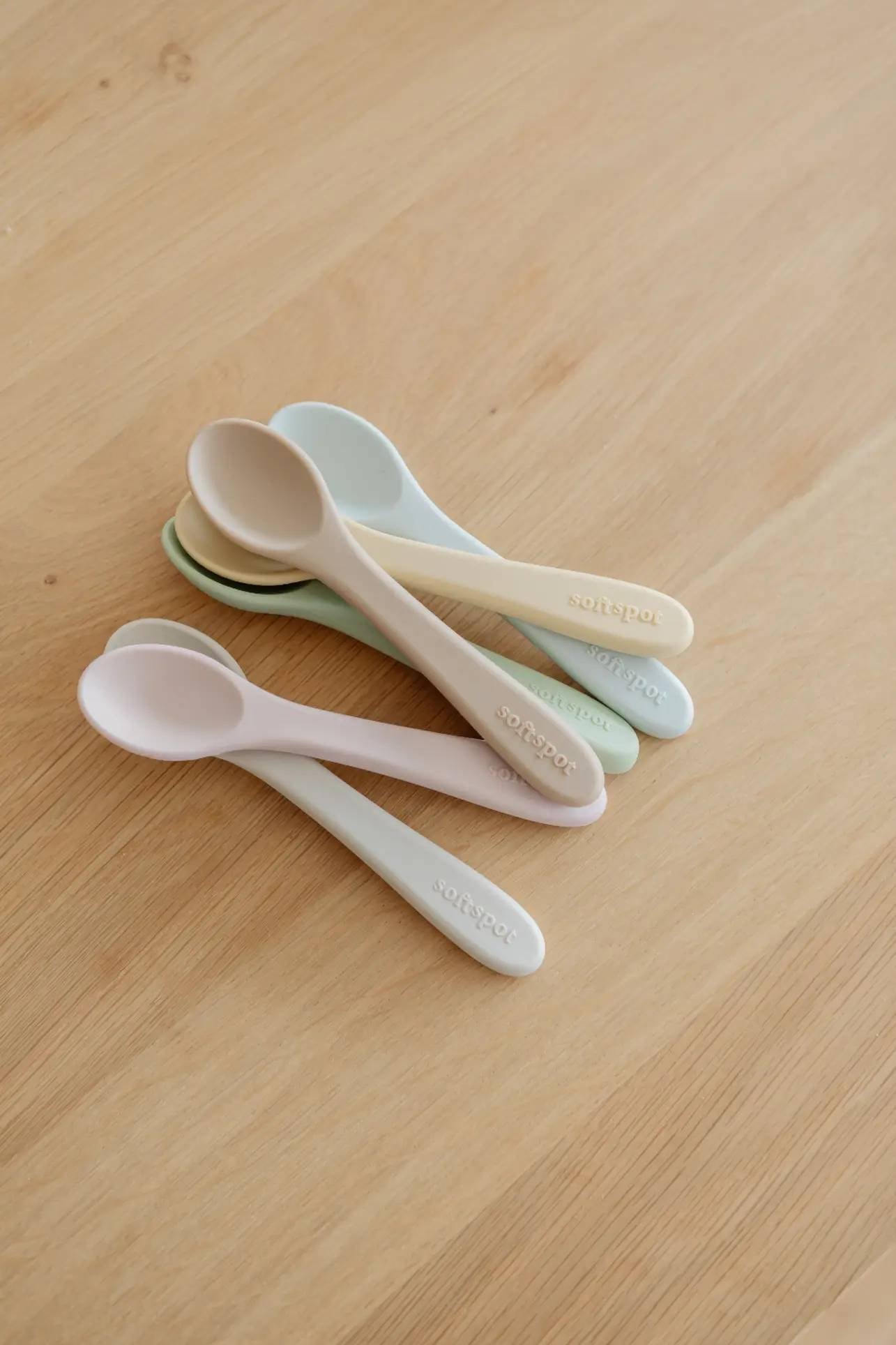 Two sets of Soft Spot Soft Baby Spoons, each in pastel shades, are stacked on a light wooden surface. Gentle on teething gums and perfect for first feedings, these spoons feature “softspot” printed on the handles.