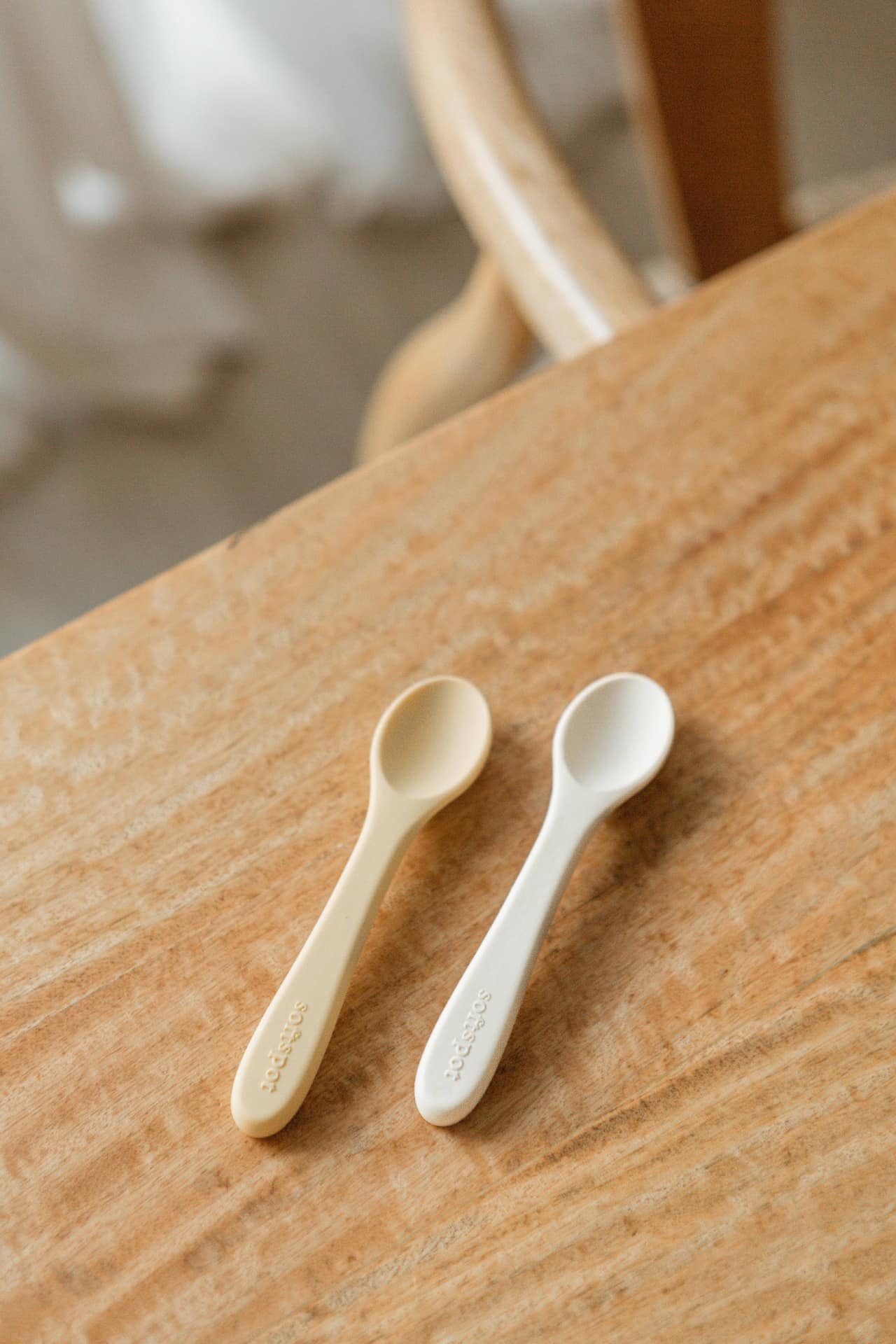 The Soft Spot Soft Baby Spoons, Set of 2 by Soft Spot, in beige and white, designed for easy feeding and gentle on gums, are placed side by side on a light wooden table with a light brown chair behind them.