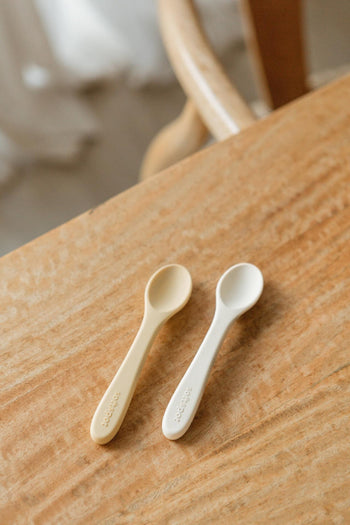 The Soft Spot Soft Baby Spoons, Set of 2 by Soft Spot, in beige and white, designed for easy feeding and gentle on gums, are placed side by side on a light wooden table with a light brown chair behind them.