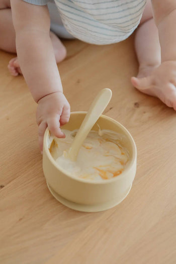A baby in a striped outfit sits on a wooden floor, reaching for the yellow Soft Spot Soft Baby Spoon from Soft Spot, perfect for easy feeding and gentle on teething gums. The matching spoon rests in a bowl of creamy food with an orange swirl.