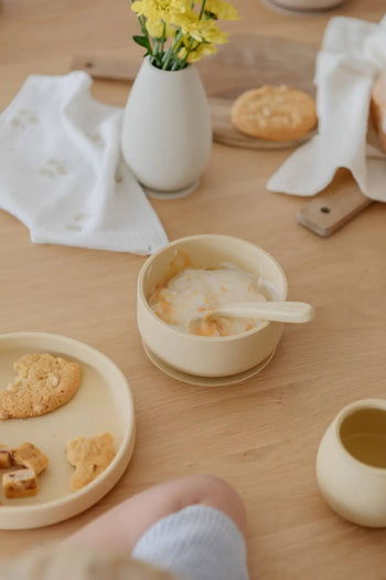 A beige bowl of yogurt with fruit and a Soft Spot Soft Baby Spoon sits on a light wooden table next to cookies, a cup, yellow flowers, and a napkin—perfect for easy feeding and soothing teething gums. Set of 2. Brand: Soft Spot.