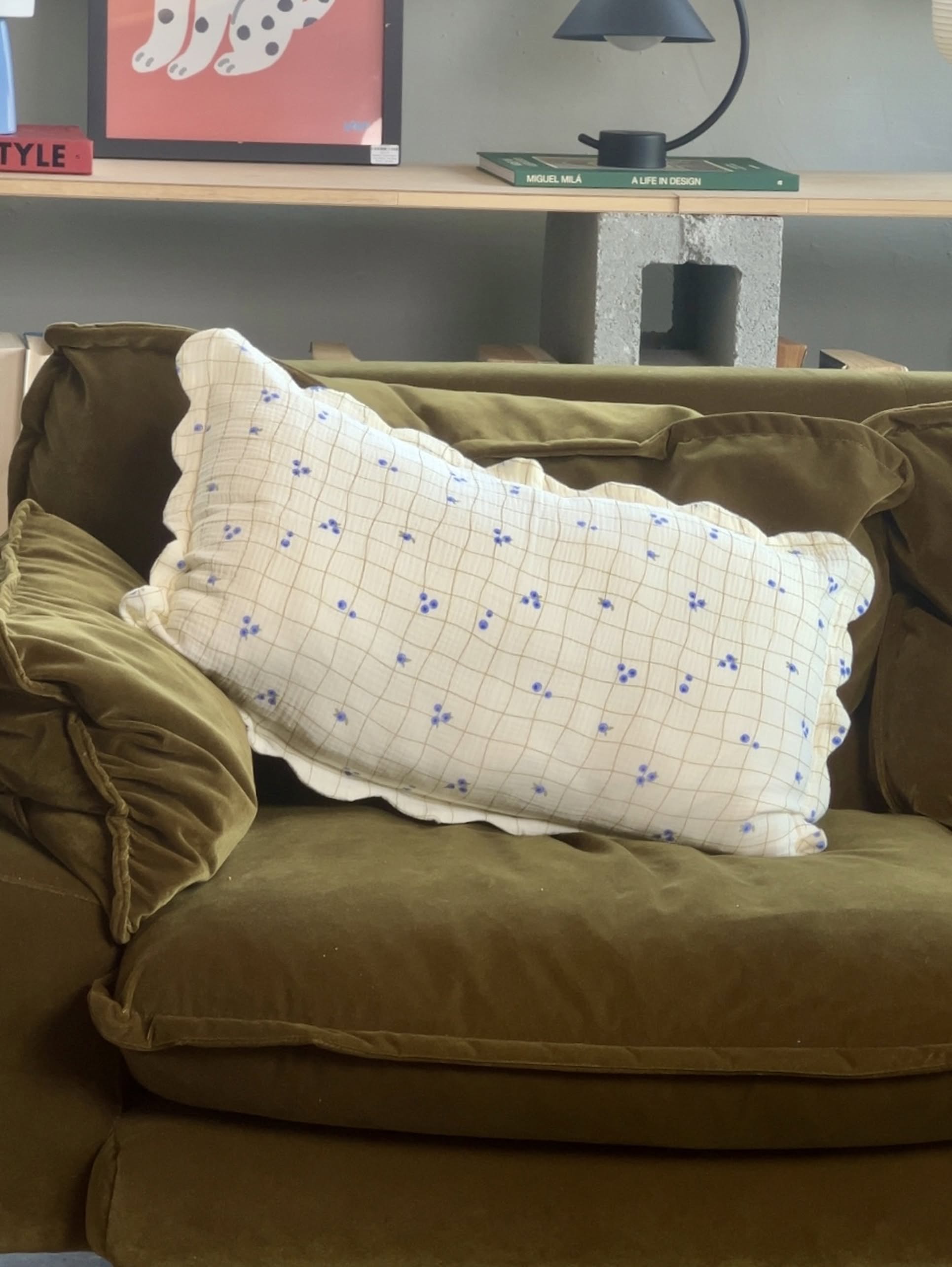 A green velvet couch with Soft Spot Soft Petal Cushion Covers Rectangle by Soft Spot. The white cushion covers feature a blue floral grid pattern. Behind the couch are shelves with books, a lamp, and colorful artwork.