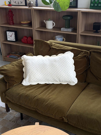 A green velvet sofa is adorned with a Soft Spot Soft Petal Cushion Cover in white, rectangular shape, while wooden shelves behind hold clocks, books, and decorative objects. In the foreground, a round wooden table completes the cozy setting.
