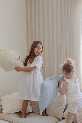 Two young girls laugh and toss Soft Spot Soft Petal Cushion Covers, Set of 2, made from light-colored fabric, on a beige sofa in a bright room with cream-colored curtains.