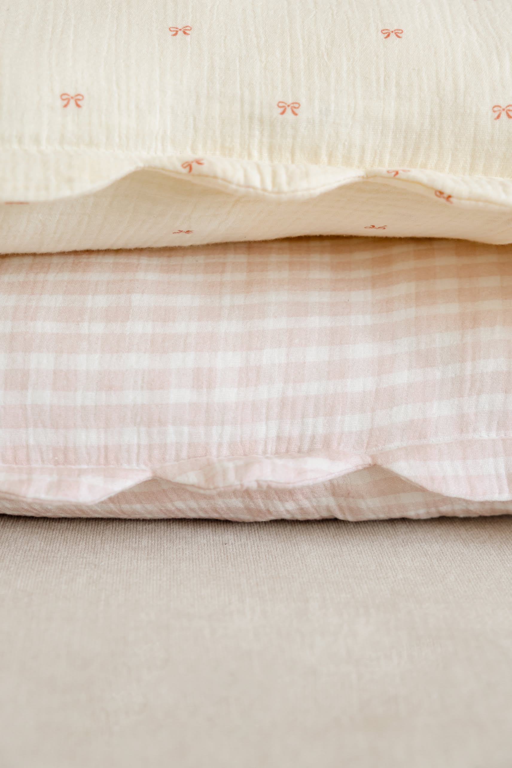 Two neatly stacked pillows, both in Soft Spot’s Soft Petal Cushion Covers (Set of 2)—one cream with pink bows on top, and one light pink with white gingham below—rest on a beige fabric surface.