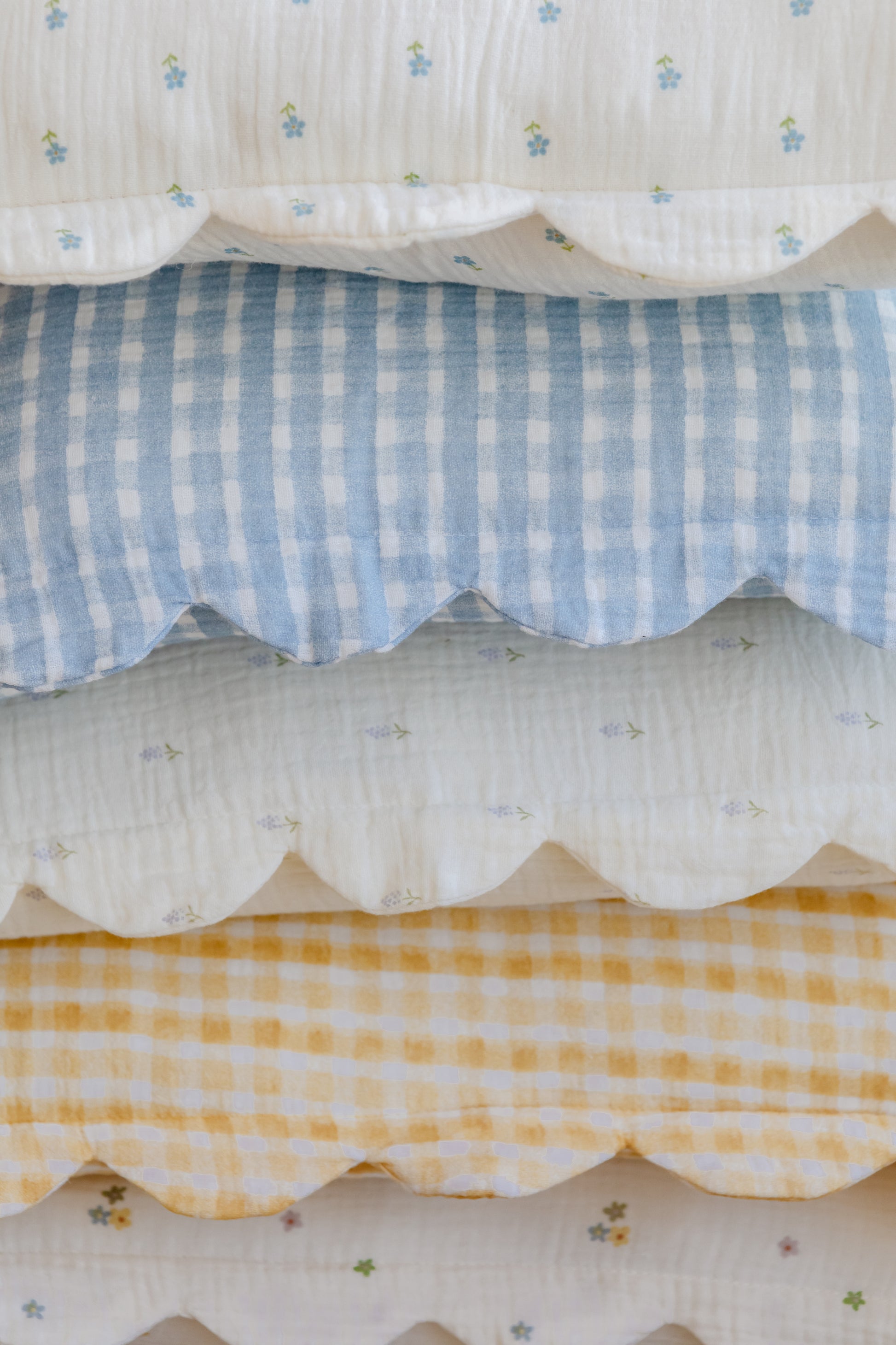 A closeup of neatly stacked bedding with scalloped edges, blue and yellow gingham, floral prints on white, and Soft Spot Soft Petal Cushion Covers (Set of 2) adding a charming finishing touch.