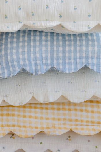 A closeup of neatly stacked bedding with scalloped edges, blue and yellow gingham, floral prints on white, and Soft Spot Soft Petal Cushion Covers (Set of 2) adding a charming finishing touch.
