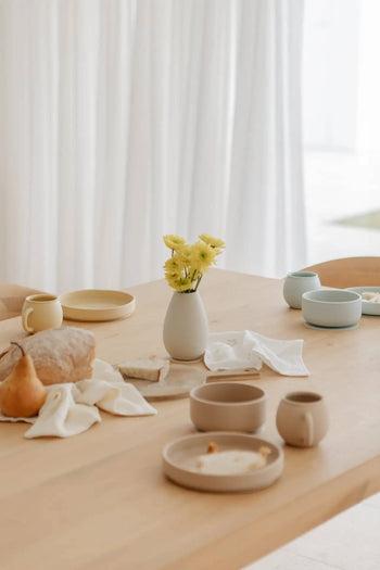A light wooden table set with neutral-toned dishes, a loaf of bread, a pear, Soft Spot Soft Piccolo Cups (Set of 2), and a small vase of yellow flowers creates an inviting mealtime scene with white curtains in the background.