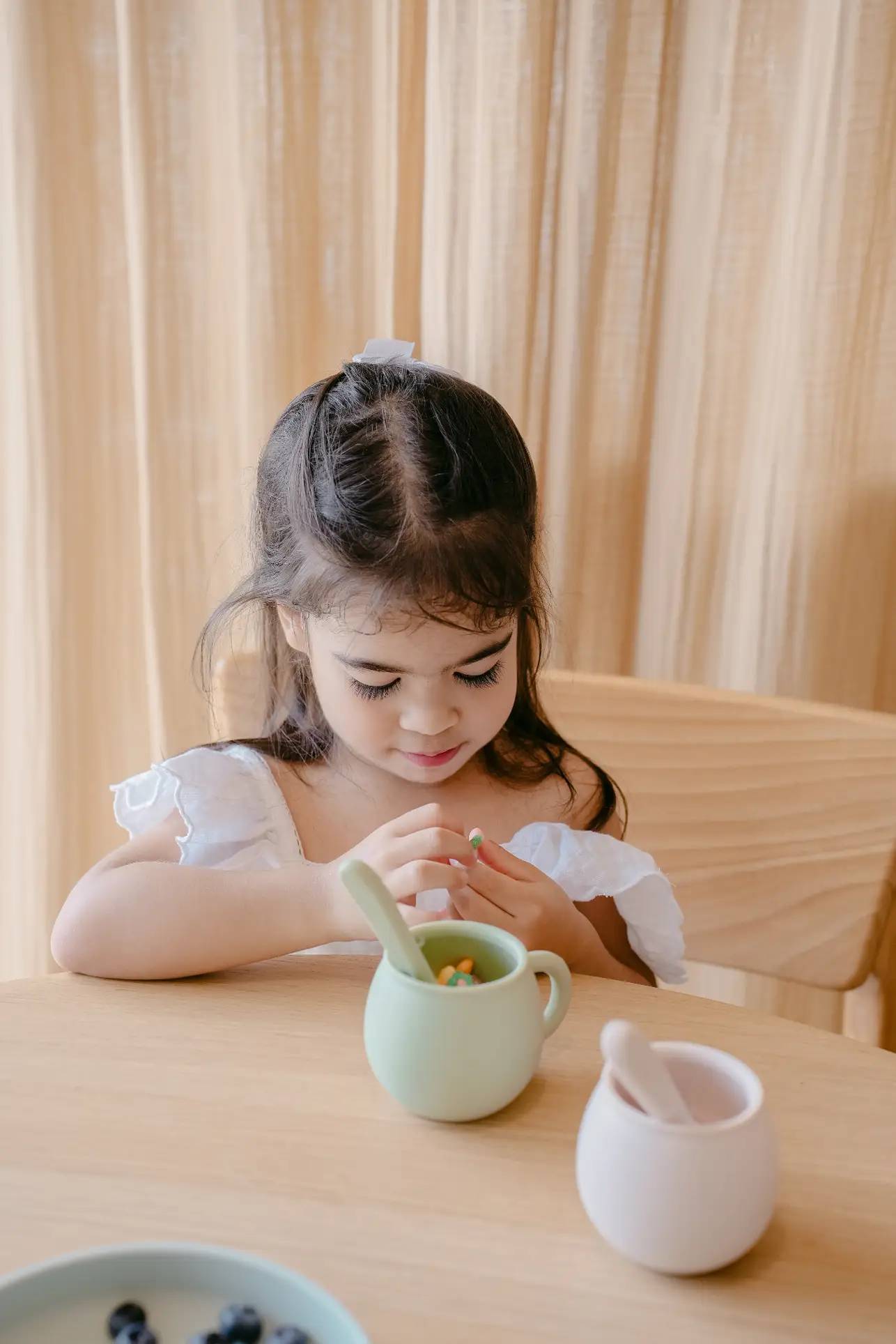 A young girl with long dark hair sits at a wooden table, where Soft Spot Soft Piccolo Cups, Set of 2, and a white mortar and pestle rest in front of her, creating a gentle mealtime scene against beige curtains.