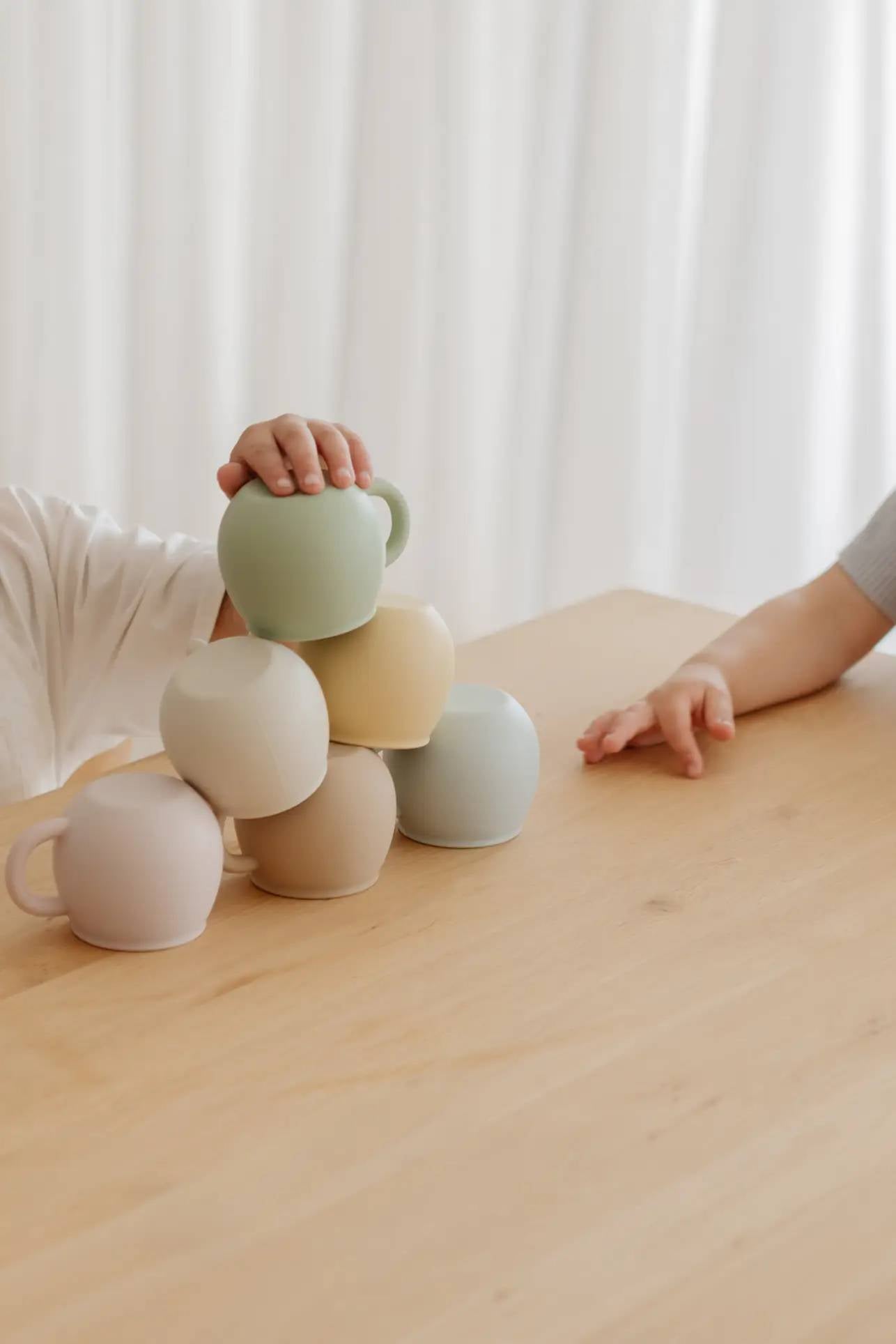 Two children stack Soft Spot Soft Piccolo Cups, Set of 2, in pastel shades into a pyramid on a light wooden table. A white curtain softly frames the background, adding a playful touch to their mealtime.