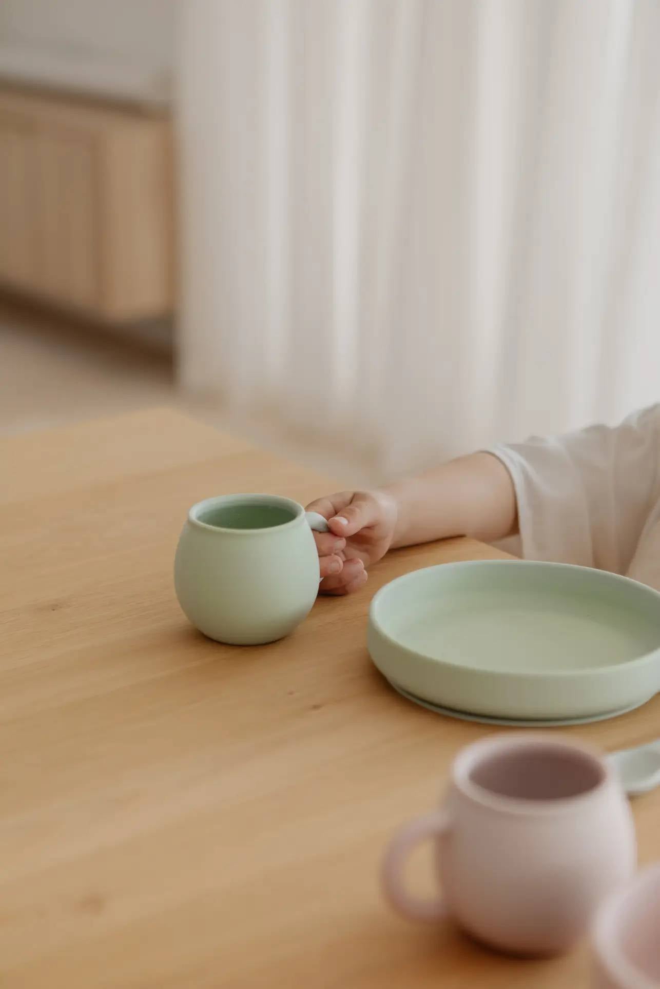 A child’s hand holds a light green Soft Spot Soft Piccolo Cup from the Soft Spot Soft Piccolo Cups, Set of 2, on a wooden table beside a matching plate, with a pink cup in the foreground and softly blurred light-colored decor in the background.
