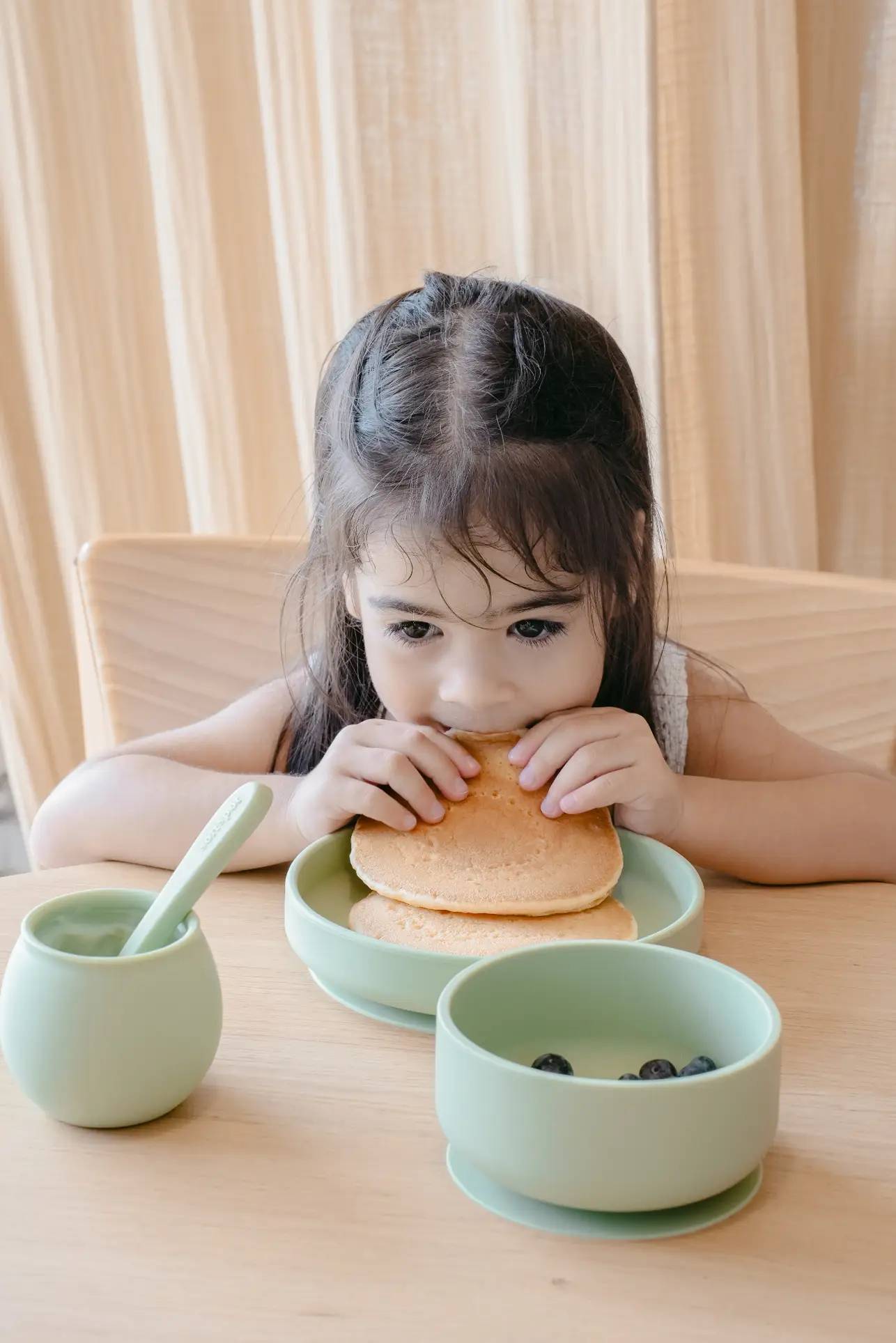 A young girl with long dark hair enjoys pancakes at a wooden table, using Soft Spot Soft Piccolo Cups, Set of 2—one filled with blueberries, the other topped with a lid and spoon. A beige curtain hangs in the background.