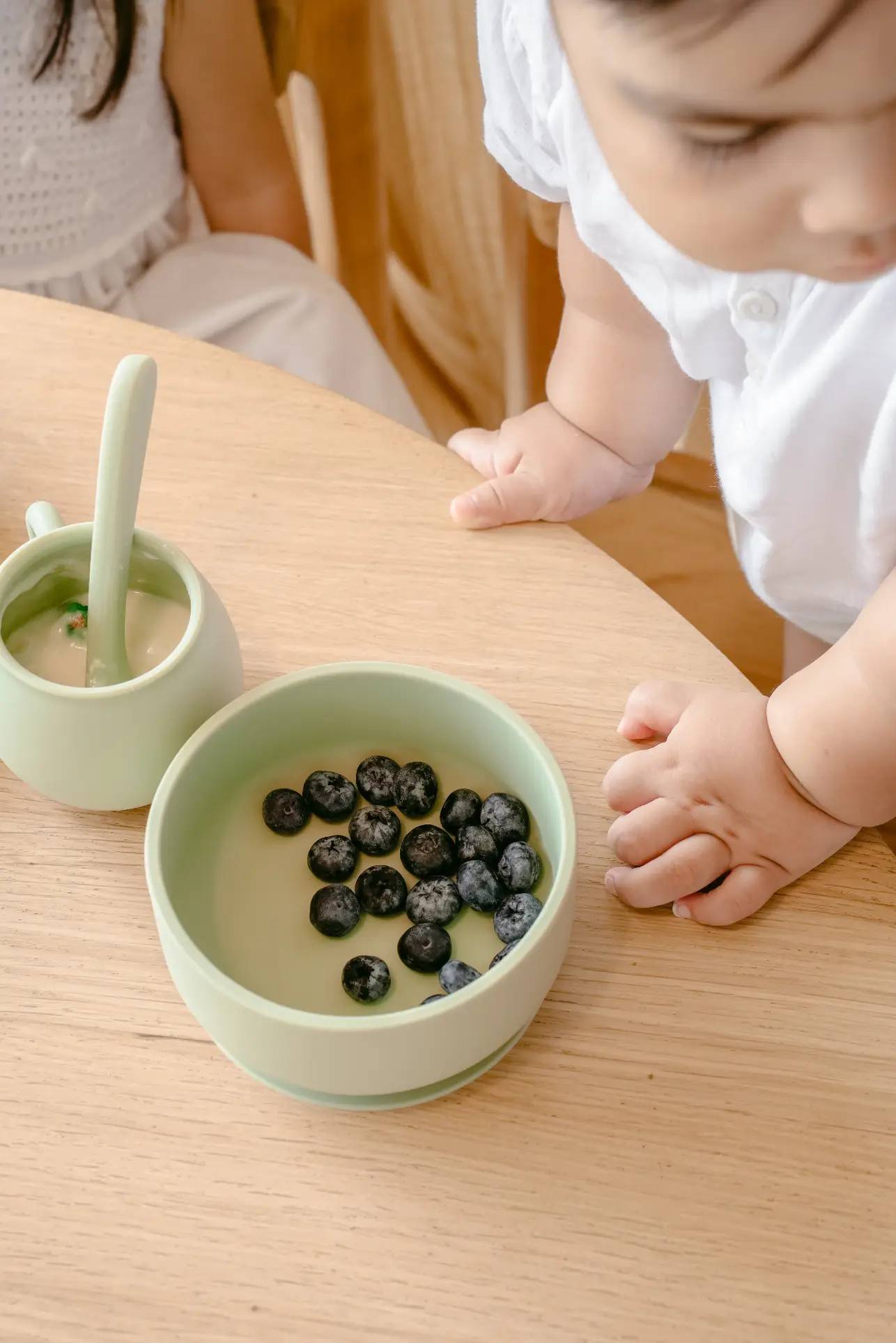 A young child in white reaches for a green bowl of blueberries and Soft Spot Soft Piccolo Cups, Set of 2—perfect baby cups—on a wooden table during mealtime. Another child appears partially in the background.