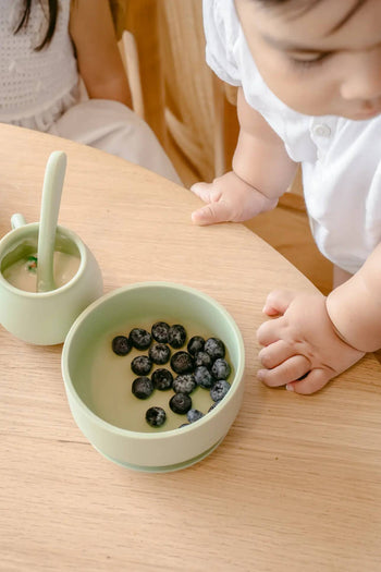 A young child in white reaches for a green bowl of blueberries and Soft Spot Soft Piccolo Cups, Set of 2—perfect baby cups—on a wooden table during mealtime. Another child appears partially in the background.