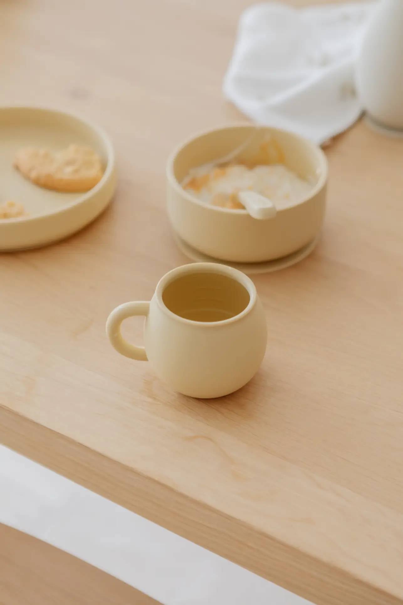 A Soft Spot Soft Piccolo Cup from the Set of 2 in beige sits on a light wooden table, next to a matching bowl with a spoon and a plate with a partially eaten cookie. A white cloth lies in the background, adding to the cozy scene.