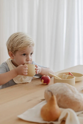 A young child with blond hair drinks from a Soft Spot Soft Piccolo Cup, part of the Soft Spot Soft Piccolo Cups Set of 2, at a table set for mealtime with a bowl, bitten apple, bread, and pear; soft white curtains frame the background.