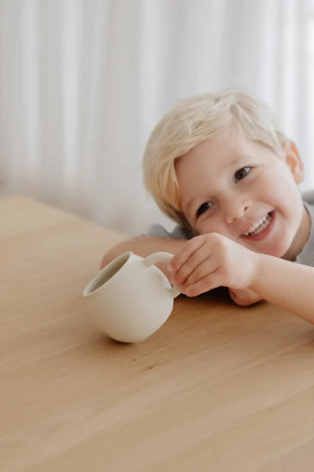 A smiling blonde child leans on a wooden table, holding a Soft Spot Soft Piccolo Cup. Light curtains in the background add to the cozy mealtime feel. Featured: Soft Spot Soft Piccolo Cups, Set of 2.