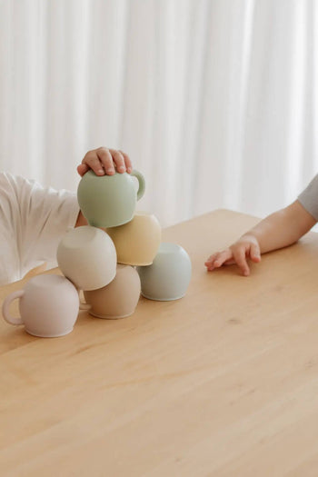 Two children’s hands stack Soft Spot Soft Piccolo Cups, Set of 2 in pastel hues on a light wooden table—perfect baby cups for mealtime, with a white curtain softly framing the playful scene.
