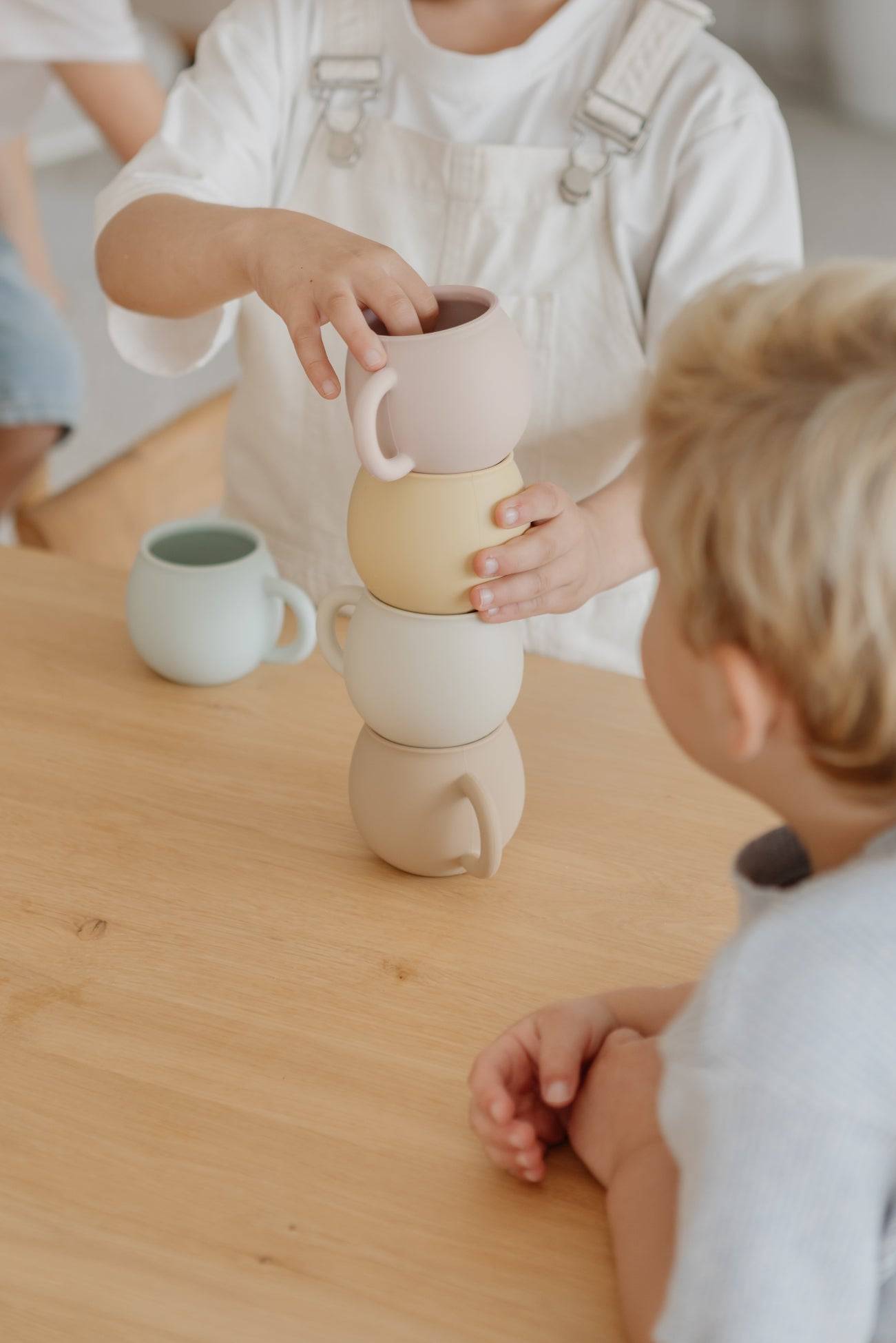 Two children sit at a wooden table. One stacks four pastel Soft Spot Soft Piccolo Cups from the Set of 2 by Soft Spot, while the other watches. Another baby cup sits nearby, highlighting their hands and a joyful mealtime moment.