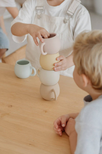 Two children sit at a wooden table. One stacks four pastel Soft Spot Soft Piccolo Cups from the Set of 2 by Soft Spot, while the other watches. Another baby cup sits nearby, highlighting their hands and a joyful mealtime moment.