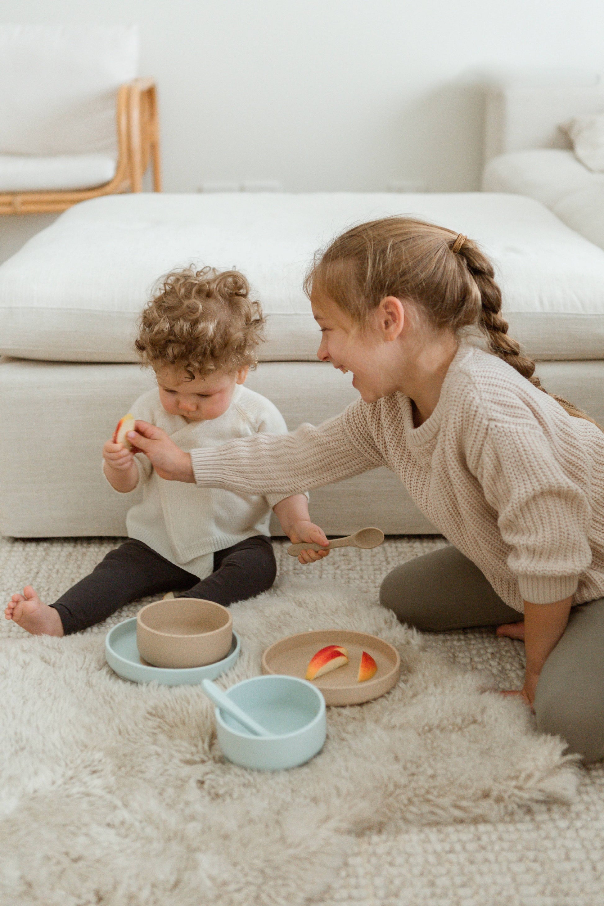 A young girl smiles as she hands fruit to a baby on a soft rug, surrounded by Soft Spot Soft Suction Bowls filled with snacks in a cozy living room.