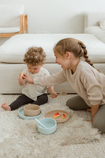 A young girl smiles as she hands fruit to a baby on a soft rug, surrounded by Soft Spot Soft Suction Bowls filled with snacks in a cozy living room.