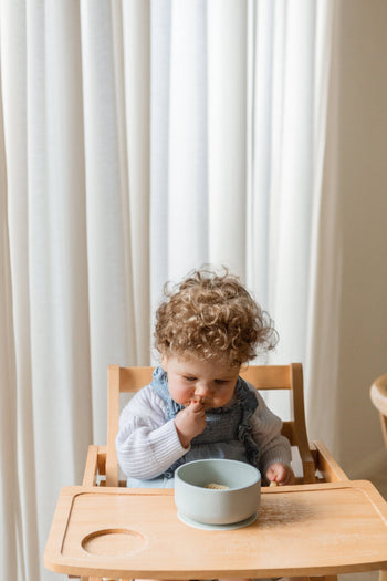 A curly-haired toddler in a light sweater and overalls enjoys a meal from the Soft Spot Soft Suction Bowl by Soft Spot, a light blue, food-grade silicone bowl with suction grip, while sitting in a wooden high chair with white curtains behind.