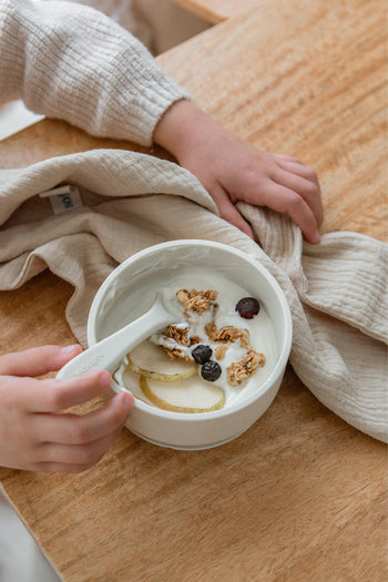 A child’s hand grips a Soft Spot Soft Baby Spoon in a bowl of yogurt with granola, apple slices, and berries on a wooden table—Soft Spot’s gentle set is perfect for little eaters and teething gums.