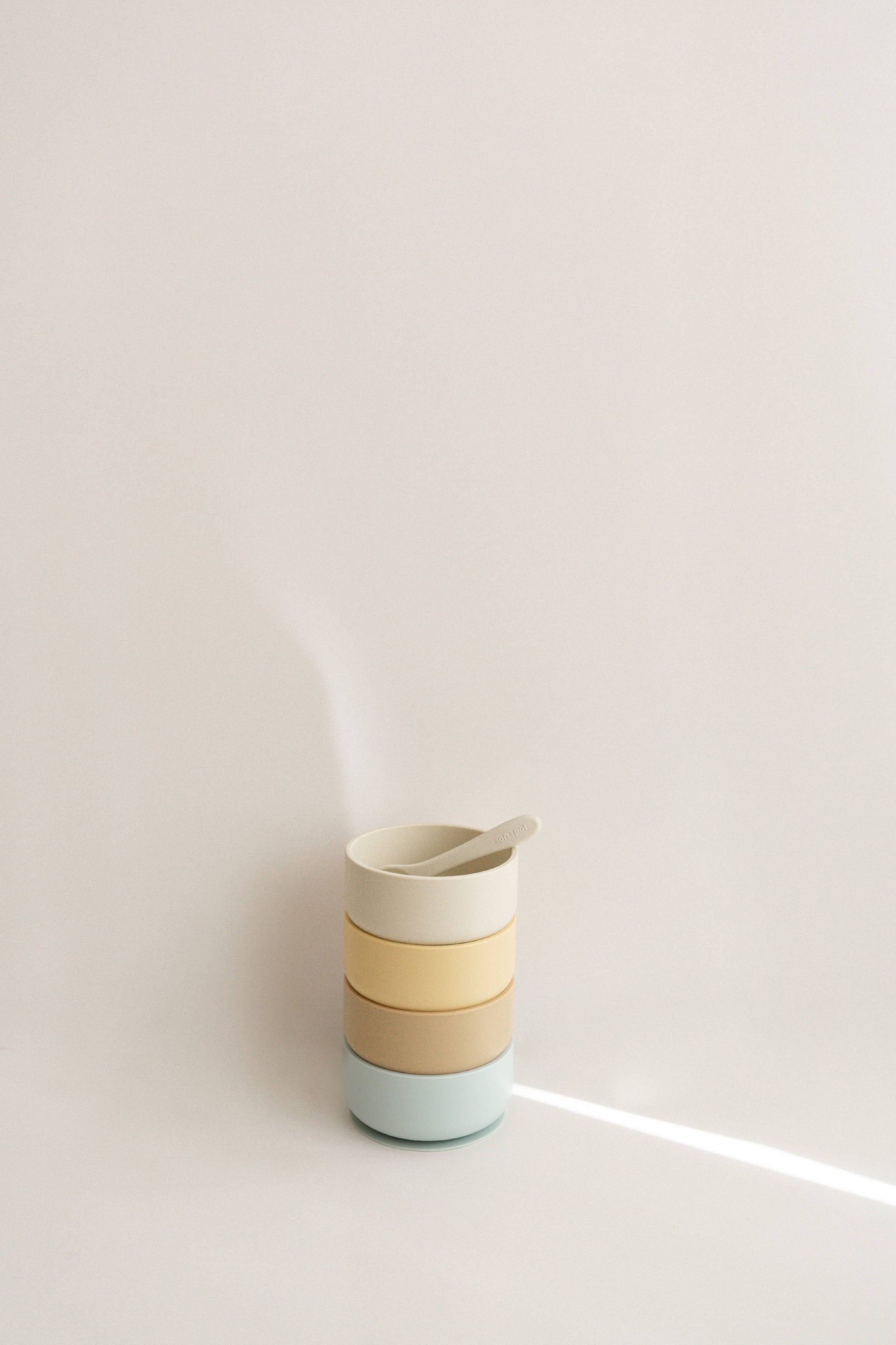 A stack of three pastel Soft Spot Soft Suction Bowls by Soft Spot, made from food-grade silicone, with a matching spoon inside, set against a plain light background with a streak of light.