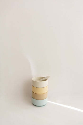 A stack of three pastel Soft Spot Soft Suction Bowls by Soft Spot, made from food-grade silicone, with a matching spoon inside, set against a plain light background with a streak of light.