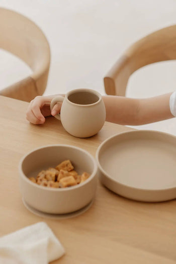 A child holds a beige mug at a wooden table, beside the Soft Spot Soft Suction Plate by Soft Spot. The food-grade silicone plate in neutral tones has a strong suction grip for secure, worry-free meals. Two wooden chairs are visible in the background.