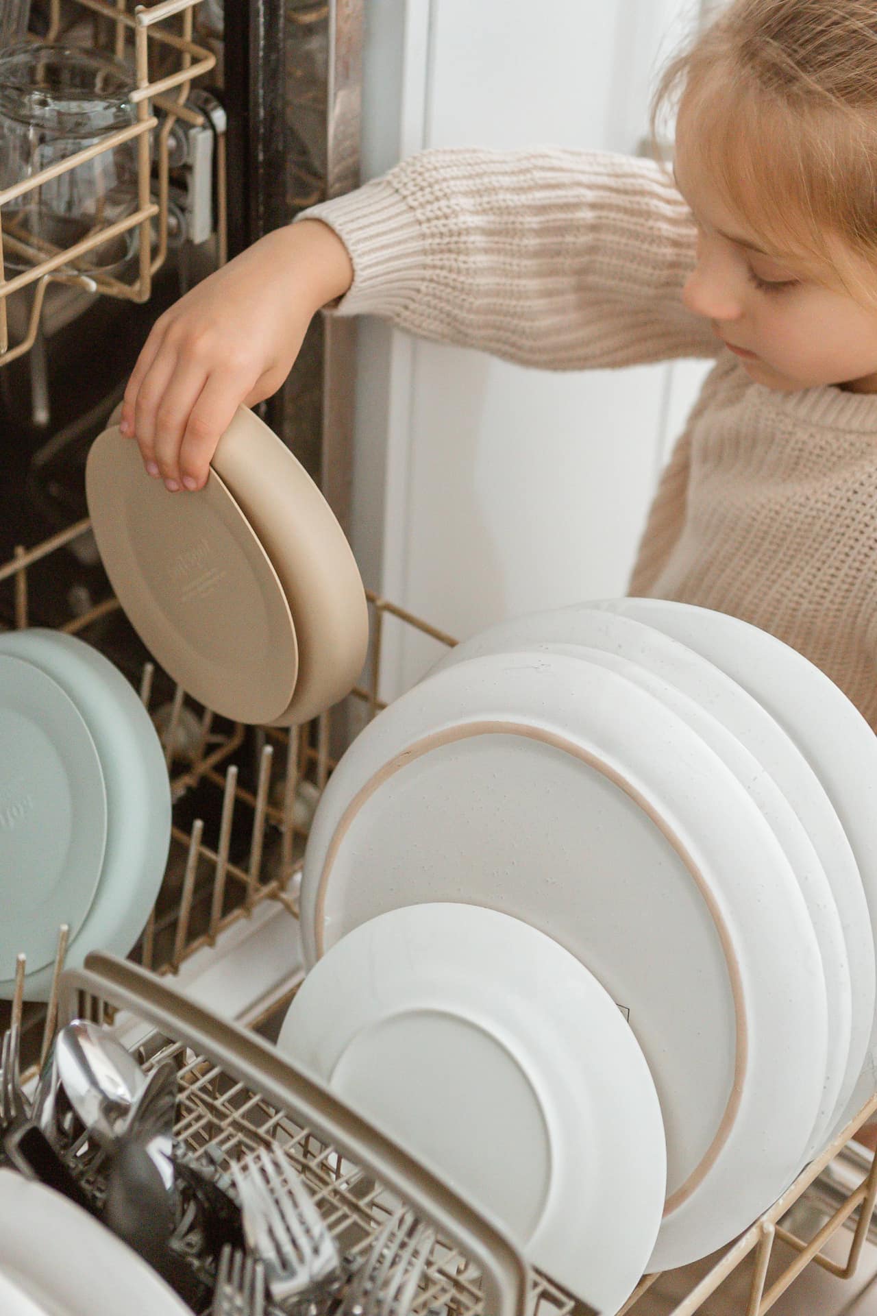 A young child in a beige sweater places a Soft Spot Soft Suction Plate by Soft Spot, made of tan food-grade silicone with strong suction grip, onto the dishwasher’s bottom rack filled with white dishes and cutlery.
