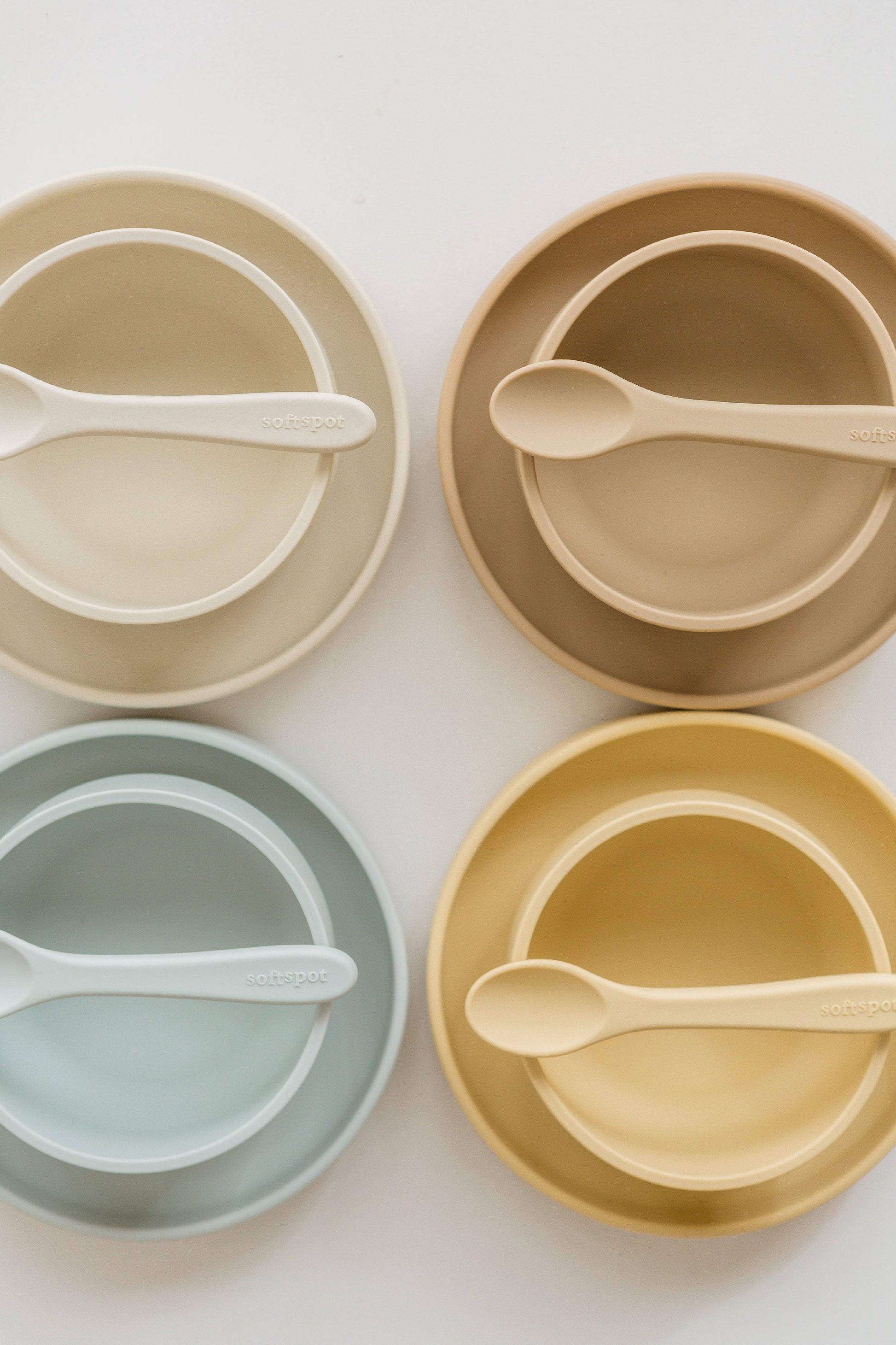 Four sets of Soft Spot Soft Suction Plates and spoons in cream, tan, light blue, and yellow are arranged in a grid on a white background. Made from food-grade silicone with strong suction grip; each spoon sits inside its matching plate.