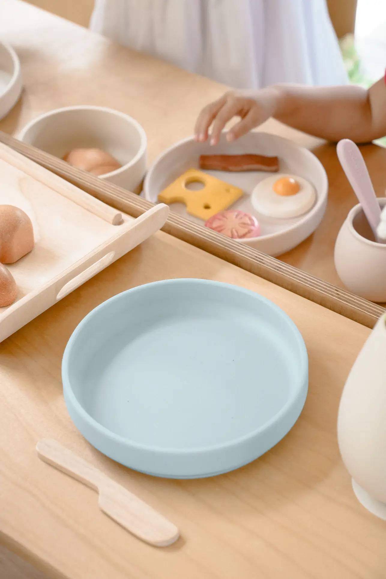 A childs hand reaches for toy food on a white food-grade silicone plate, next to a blue Soft Spot Soft Suction Plate by Soft Spot, a wooden knife, and other toy dishes on a light wooden table.
