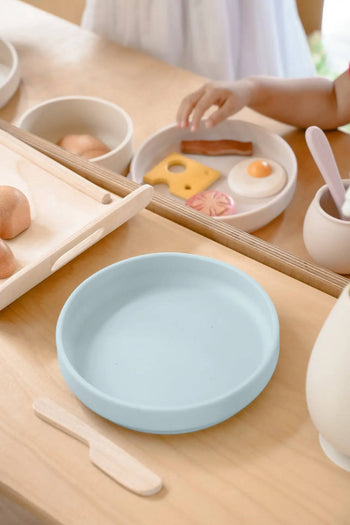 A childs hand reaches for toy food on a white food-grade silicone plate, next to a blue Soft Spot Soft Suction Plate by Soft Spot, a wooden knife, and other toy dishes on a light wooden table.