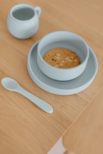 A Soft Spot Soft Suction Plate by Soft Spot in light blue holds a cookie on a wooden table, with a matching baby spoon and small cup nearby.
