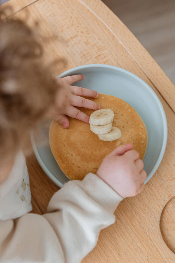 A young child reaches for pancakes with banana slices on the Soft Spot Soft Suction Plate by Soft Spot, a food-grade silicone plate with strong suction grip, set on a wooden table.