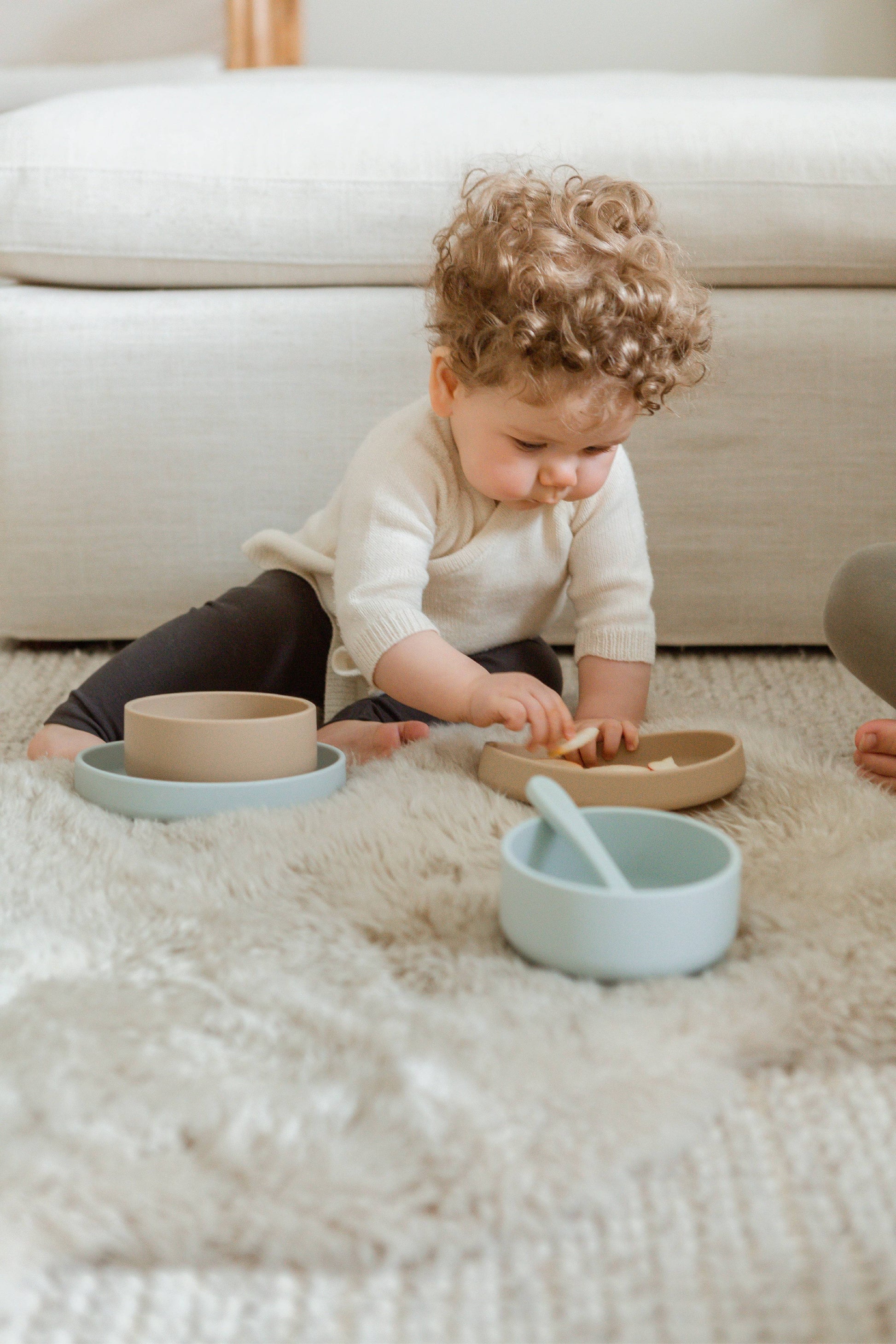 A young child with curly hair sits on a rug, playing with beige and blue Soft Spot Soft Suction Plates from Soft Spot, made of food-grade silicone for strong suction. A couch is visible in the background.