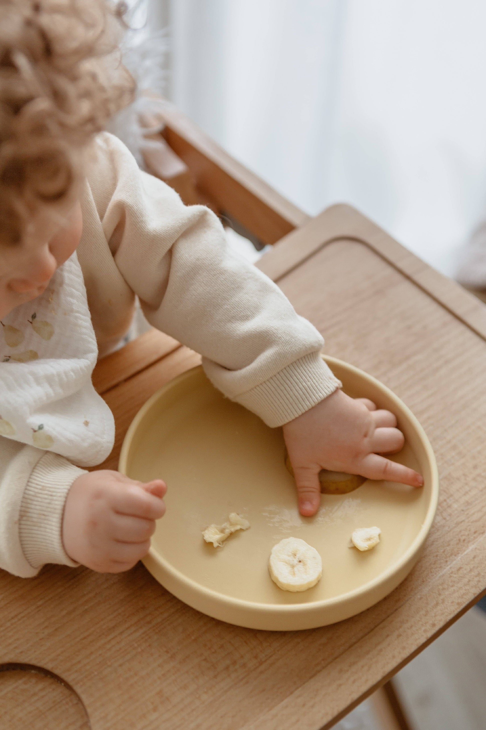 A young child sits in a wooden high chair, reaching for banana slices on the Soft Spot Soft Suction Plate by Soft Spot. The yellow food-grade silicone plate features a strong suction grip to stay securely in place.