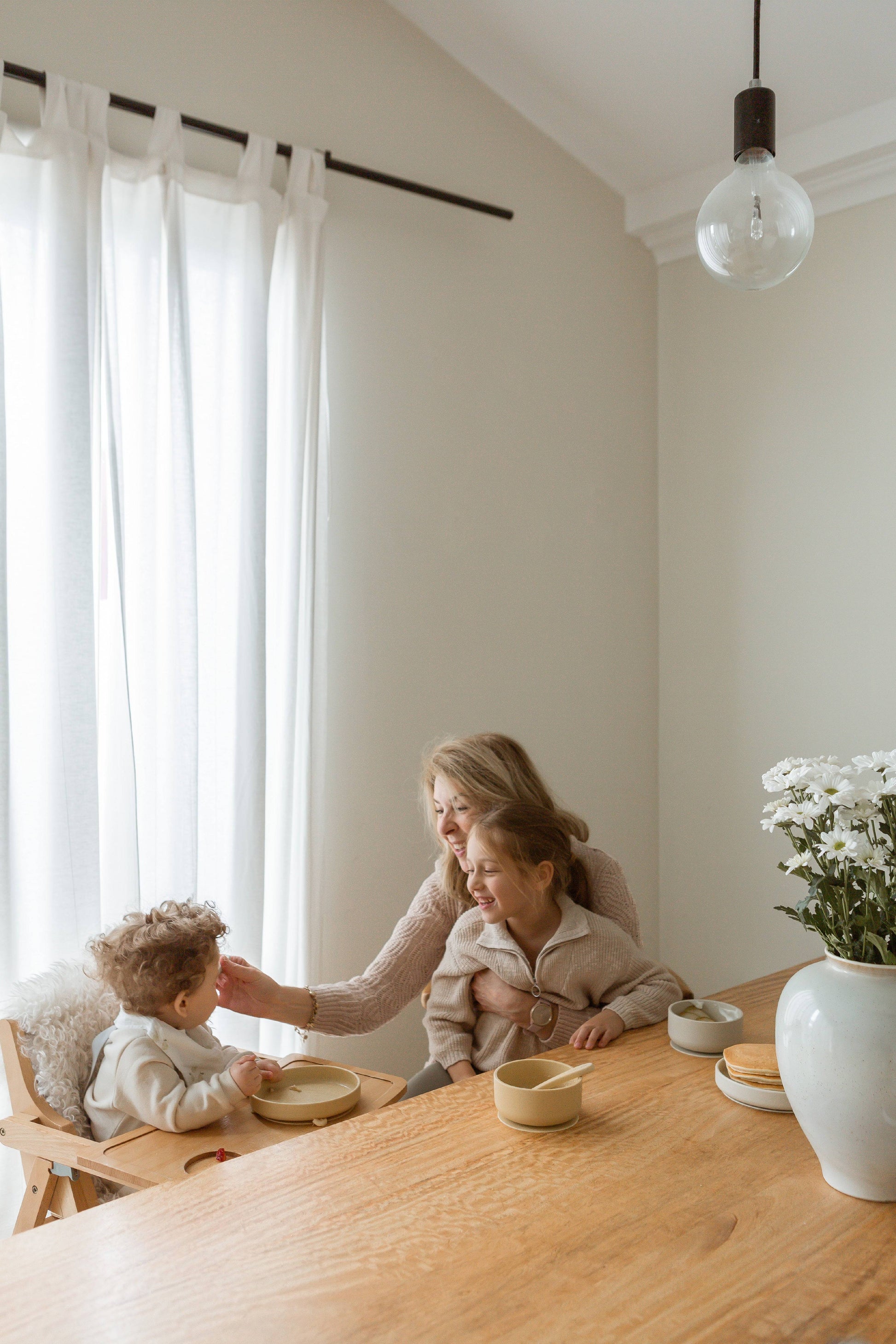 A woman sits at a wooden table with two young children, gently feeding the younger child from a Soft Spot Soft Suction Plate by Soft Spot. Sunlight filters through white curtains, while a vase of white flowers adds to the warm, peaceful atmosphere.