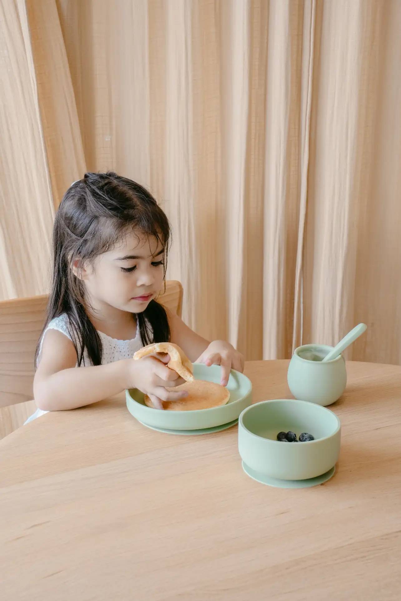 A young girl with long dark hair sits at a wooden table, picking up a pancake from the Soft Spot Soft Suction Plate by Soft Spot. Beside her are a green cup with a spoon and a bowl of blueberries, with light curtains in the background.