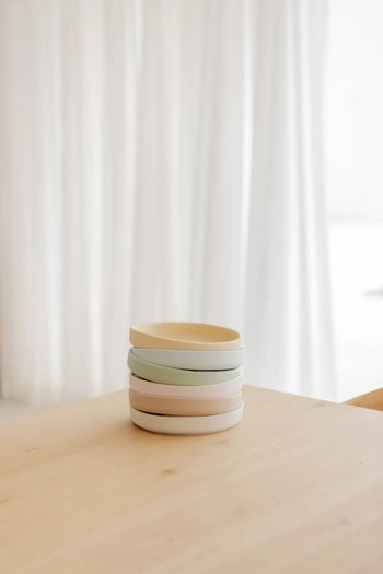 The Soft Spot Soft Suction Plate by Soft Spot, made from food-grade silicone in neutral tones, sits securely on a light wooden table with its strong suction grip, as sheer white curtains glow softly in the background.