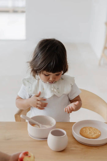 A young child with dark hair sits at a wooden table, gazing at their hand. In front of them are a Soft Spot Soft Suction Plate by Soft Spot with a cookie on it and a cup. The scene is minimal and softly lit.
