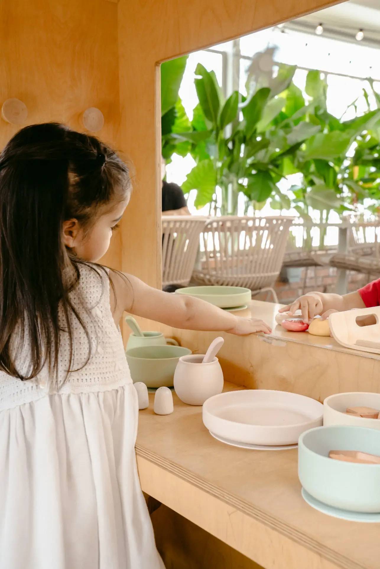 A young girl in a white dress plays with the Soft Spot Soft Suction Plate by Soft Spot on a wooden counter, while a mirror reflects her actions and green plants accent the background.