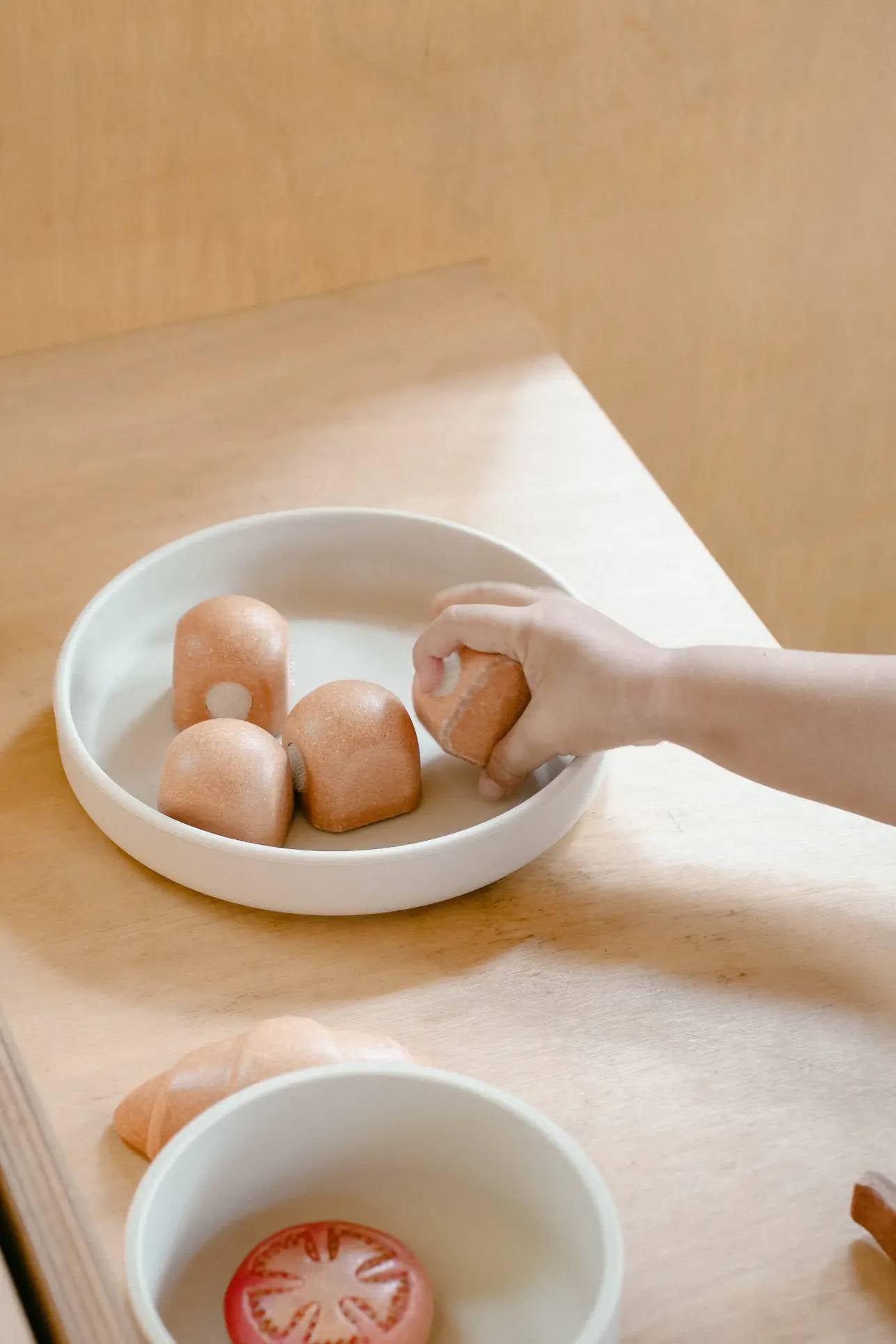 A child’s hand lifts a wooden toy bread roll from the Soft Spot Soft Suction Plate by Soft Spot on a light wooden table, with more wooden food toys like a tomato slice in another bowl nearby.